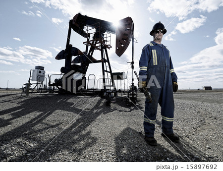 A man in overalls and hard hat at a pump jack in open ground at an oil extraction site. 15897692