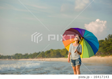 Young caucasian woman wearing sunglasses with colourful rainbow umbrella looking at ocean on the Young caucasian woman wearing sunglasses with colourful rainbow umbrella looking at ocean on the 16417143