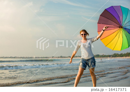 Cheerful young girl with rainbow umbrella having fun on the beach before sunset. Travel, holidays 16417371