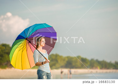 Young caucasian woman wearing sunglasses with colourful rainbow umbrella looking at ocean on the Young caucasian woman wearing sunglasses with colourful rainbow umbrella looking at ocean on the 16683130