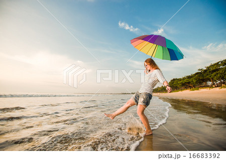 Cheerful young girl with rainbow umbrella having fun on the beach before sunset. Travel, holidays 16683392
