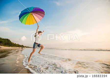 Cheerful young girl with rainbow umbrella having fun on the beach before sunset. Travel, holidays Cheerful young girl with rainbow umbrella having fun on the beach before sunset. Travel, holidays 16683394