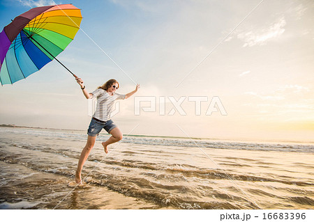 Cheerful young girl with rainbow umbrella having fun on the beach before sunset. Travel, holidays Cheerful young girl with rainbow umbrella having fun on the beach before sunset. Travel, holidays 16683396