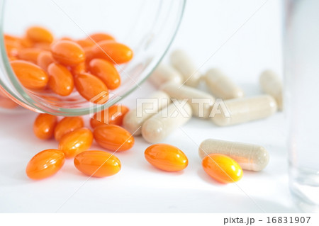 透明の瓶からこぼれ出したオレンジ色とベージュ色のサプリメントとグラスに入った水 16831907