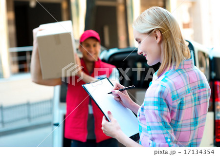 Sign here please! Smiling young delivery man holding a cardboard box while beautiful young woman 17134354