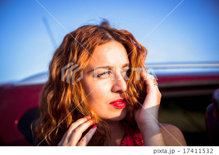 Beautiful red-haired young woman posing by headlight of Red luxury car. Attractive girl with red 17411532