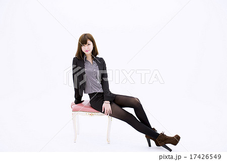 白い背景の前で椅子に横向きで座りながら両足を投げ出してポーズをとる微笑む若いスーツ姿の女性の写真素材