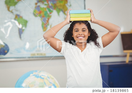 Smiling pupil balancing books on head in a classroom 18139865