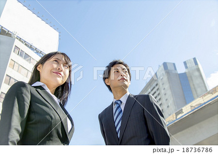 青空バックのビル群の前で遠くを見上げるスーツ姿の若い男女 18736786