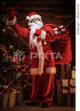 Santa Claus in wooden home interior with sack full of Christmas presents Santa Claus in wooden home interior with sack full of Christmas presents 18921235