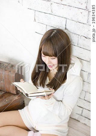 白い壁にもたれてあぐらで座り両手で本を読んでいるモコモコの部屋着の笑顔の若い女性 19634385