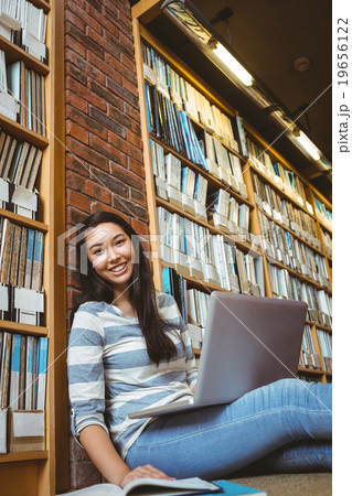 Smiling student sitting on the floor against wall in library studying with laptop and books Smiling student sitting on the floor against wall in library studying with laptop and books 19656122