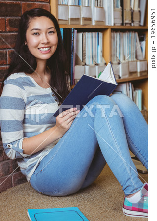 Smiling student sitting on the floor against wall in library reading book 19656951