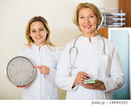 Two friendly doctors holding clock and smiling in modern clinic 19694305