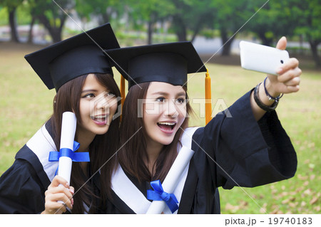 Two happy women in graduation gowns taking picture with cell pho 19740183