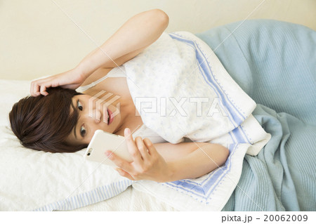 ベッドで寝ながらスマホを操作する若い女性の写真素材 0609