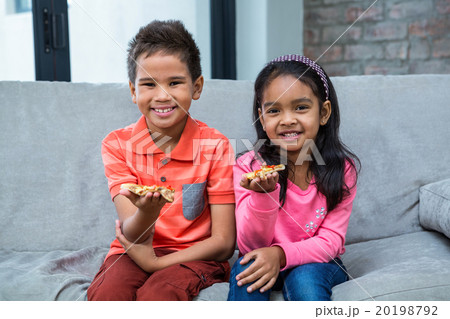 Smiling siblings eating pizza on the sofa 20198792