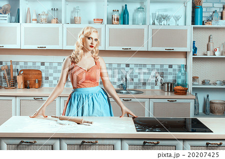 Blonde girl standing in the kitchen next to stove. 20207425