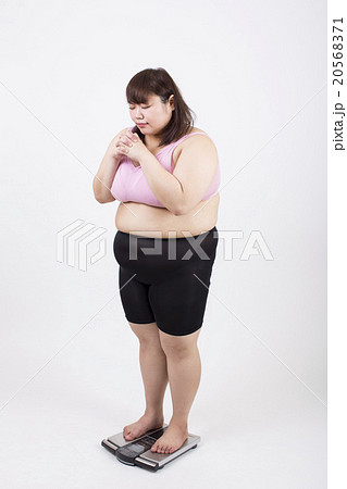白い背景の前で祈るような気持ちで体重計に乗っている太った若い女性 20568371