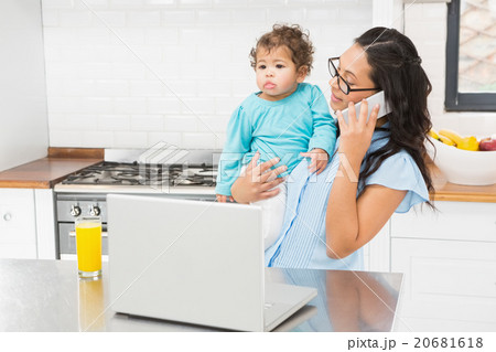 Smiling brunette holding her baby and using laptop on phone call Smiling brunette holding her baby and using laptop on phone call 20681618