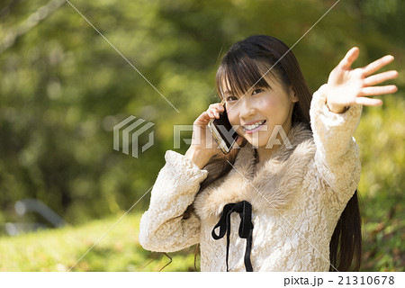 緑の森林公園で嬉しそうにスマートフォンで会話通話をしながら手を振る可愛い笑顔の女性の写真素材