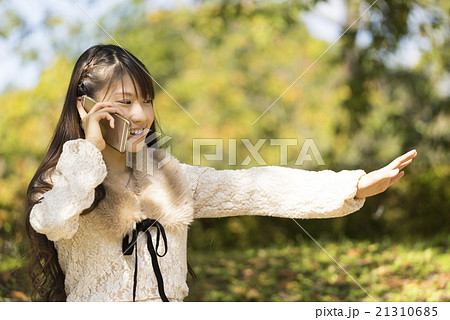 緑の森林公園で嬉しそうにスマートフォンで会話通話をすしながら手を振る可愛い笑顔の女子学生 21310685