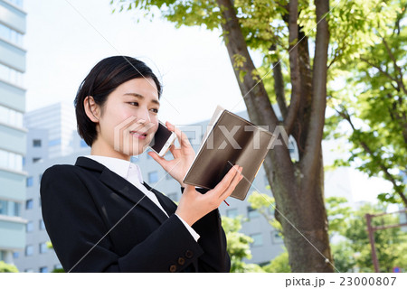 ビジネス街で手帳を確認しながら電話する若い会社員の女性 困る ビジネス街で手帳を確認しながら電話する若い会社員の女性 困る 23000807