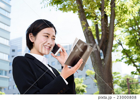 ビジネス街で手帳を確認しながら電話する若い会社員の女性 23000808
