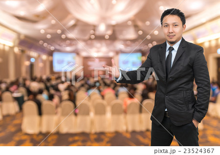 Asian Businessman with welcoming gesture on Abstract blurred photo of conference hall or seminar room with attendee background Asian Businessman with welcoming gesture on Abstract blurred photo of conference hall or seminar room with attendee background 23524367
