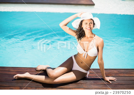 Portrait of happy woman in hat sitting on wooden decker by pool Portrait of happy woman in hat sitting on wooden decker by pool 23970250