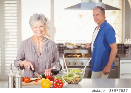 Happy senior woman cutting vegetables with husband in background Happy senior woman cutting vegetables with husband in background 23977627
