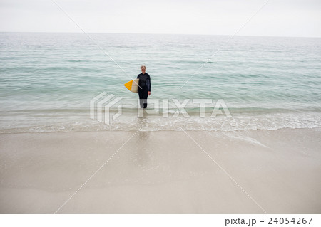 Senior woman in wetsuit standing in water with surfboard on the beach 24054267
