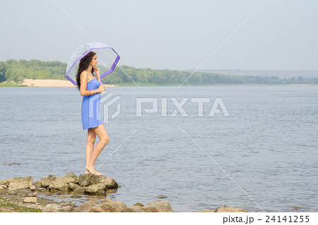 Lonely sad young girl with an umbrella stands on the bank of the river and looks into the distance 24141255