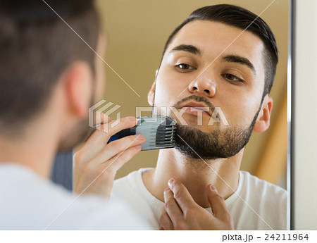 Man looking at mirror and shaving beard with trimmer 24211964