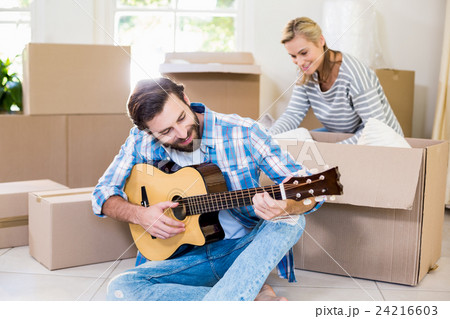 Man playing a guitar while woman unpackaging cardboard boxes in background 24216603
