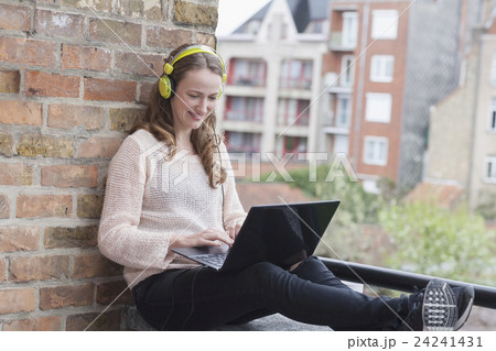 Mid-adult woman with headphones on sitting on balcony railing and using laptop 24241431