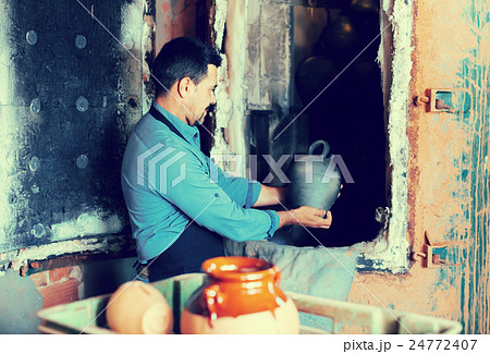 mature man potter holding black glazed ceramic vessel next to kiln 24772407