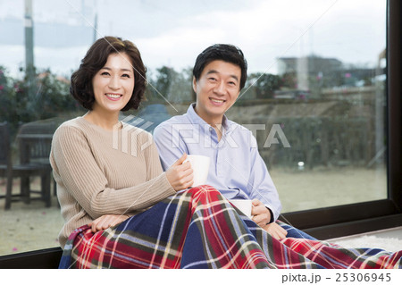 Middleaged Asian Couple Smiling and Sitting with Coffee Mugs on Rug In Livingroom 25306945