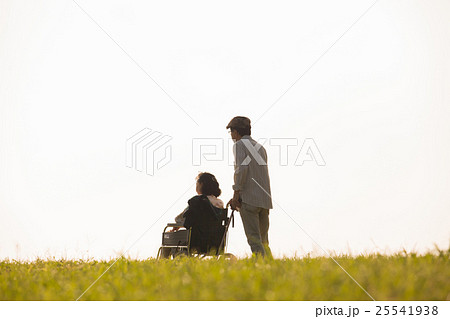 夕暮れの丘で景色を眺める車いすに乗った年老いた妻と介助する夫 25541938