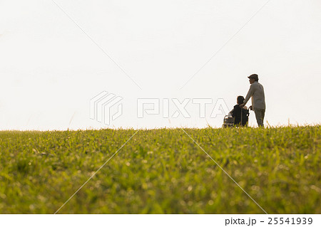 夕暮れの丘で景色を眺める車いすに乗った年老いた妻と介助する夫 25541939