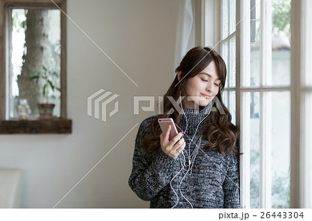 スマートフォンで音楽を聴きながら窓辺に佇む若い女性 スマートフォンで音楽を聴きながら窓辺に佇む若い女性 26443304