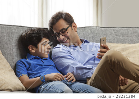 Smiling father and son sitting on sofa using mobile phone Smiling father and son sitting on sofa using mobile phone 28231284