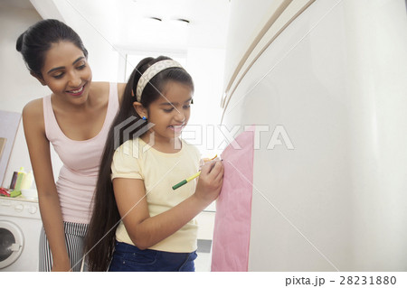 Mother looking at daughter writing note on refrigerator 28231880