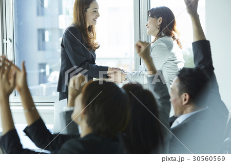 Two businesswomen are shaking hands and a group of people is cheering. 30560569