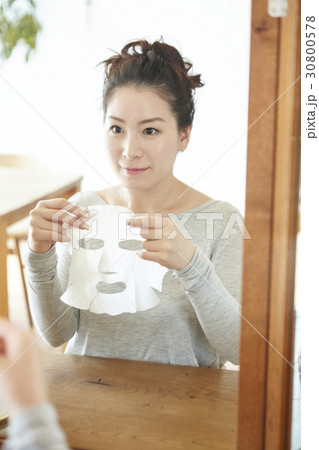 a woman sits in front of a mirror and lifts up facial mask sheet. a woman sits in front of a mirror and lifts up facial mask sheet. 30800578