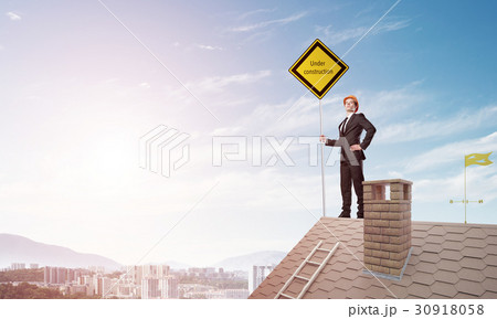 Young businessman on house brick roof holding yellow signboard and looking at city. Mixed media Young businessman on house brick roof holding yellow signboard and looking at city. Mixed media 30918058