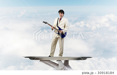 Businessman on metal tray playing electric guitar against blue sky background 30918184