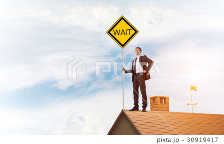 Young businessman on house brick roof holding yellow signboard. Mixed media 30919417