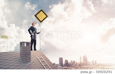 Young businessman on house brick roof holding yellow signboard a Young businessman on house brick roof holding yellow signboard a 30920291