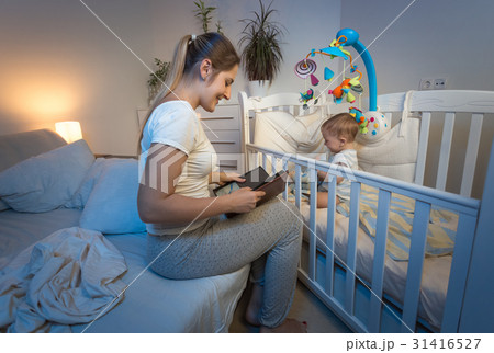 Mother sitting at baby's crib and reading a book Mother sitting at baby's crib and reading a book 31416527